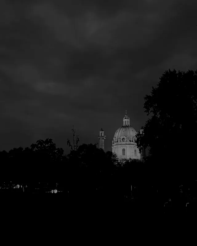 La Karlskirche de Viena iluminada de noche, vista de lejos detrás de los árboles.